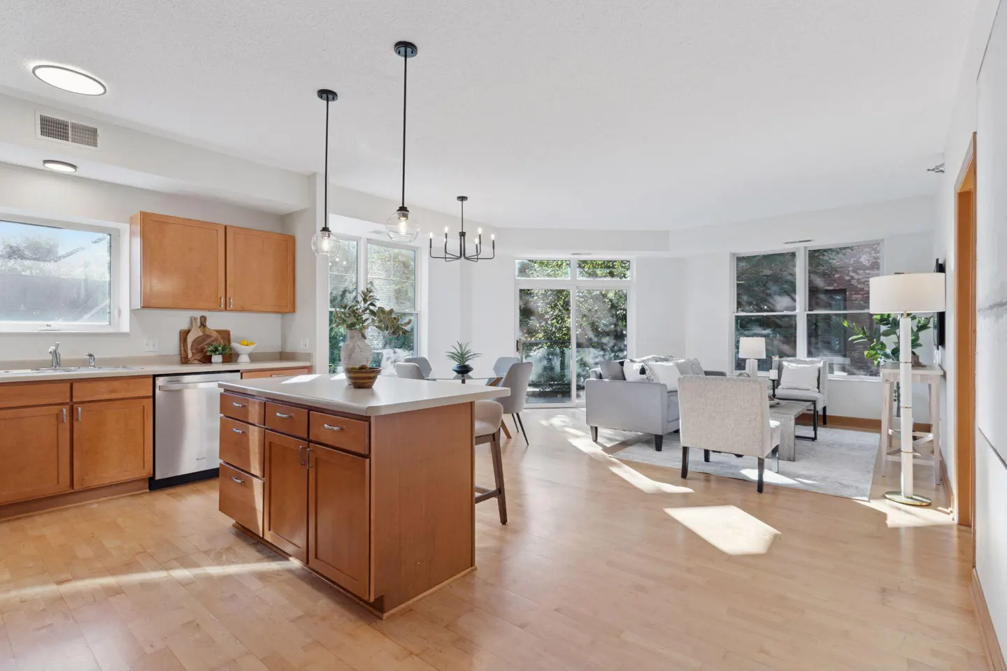 Open-concept kitchen and living room with wood floors. Kitchen island, wood cabinets, and stainless steel appliances. Living room has gray sofa and chairs.