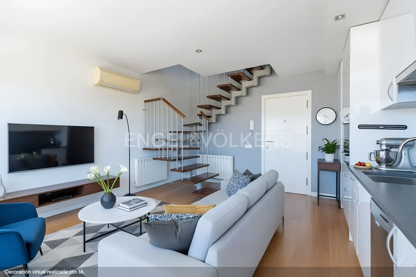 Bright, modern living room with a gray sofa, wood floors, and floating stairs. A TV and kitchen are visible.