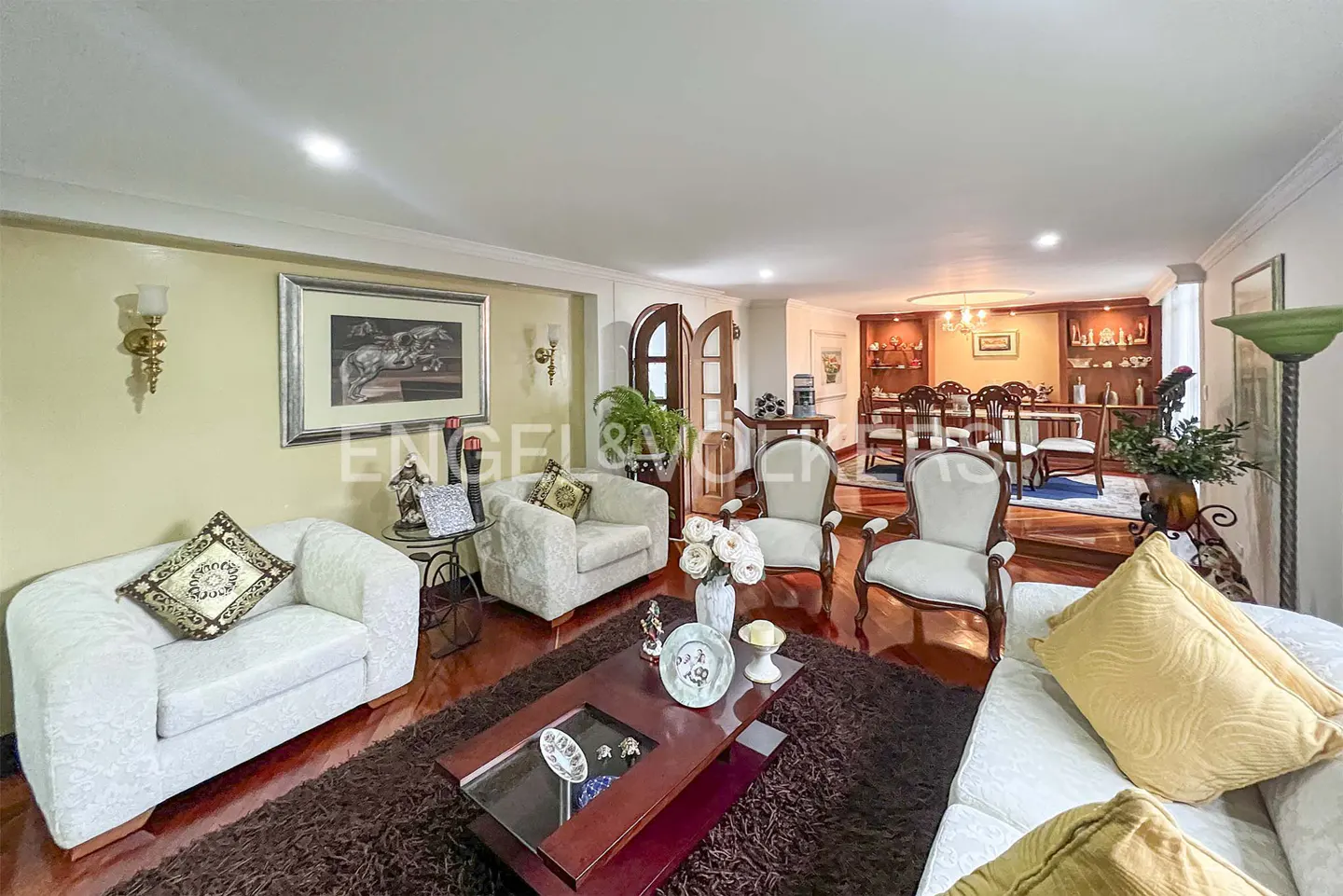 Living room with white sofas, armchairs, and a dark wood coffee table on a brown rug. Dining room visible in the background.