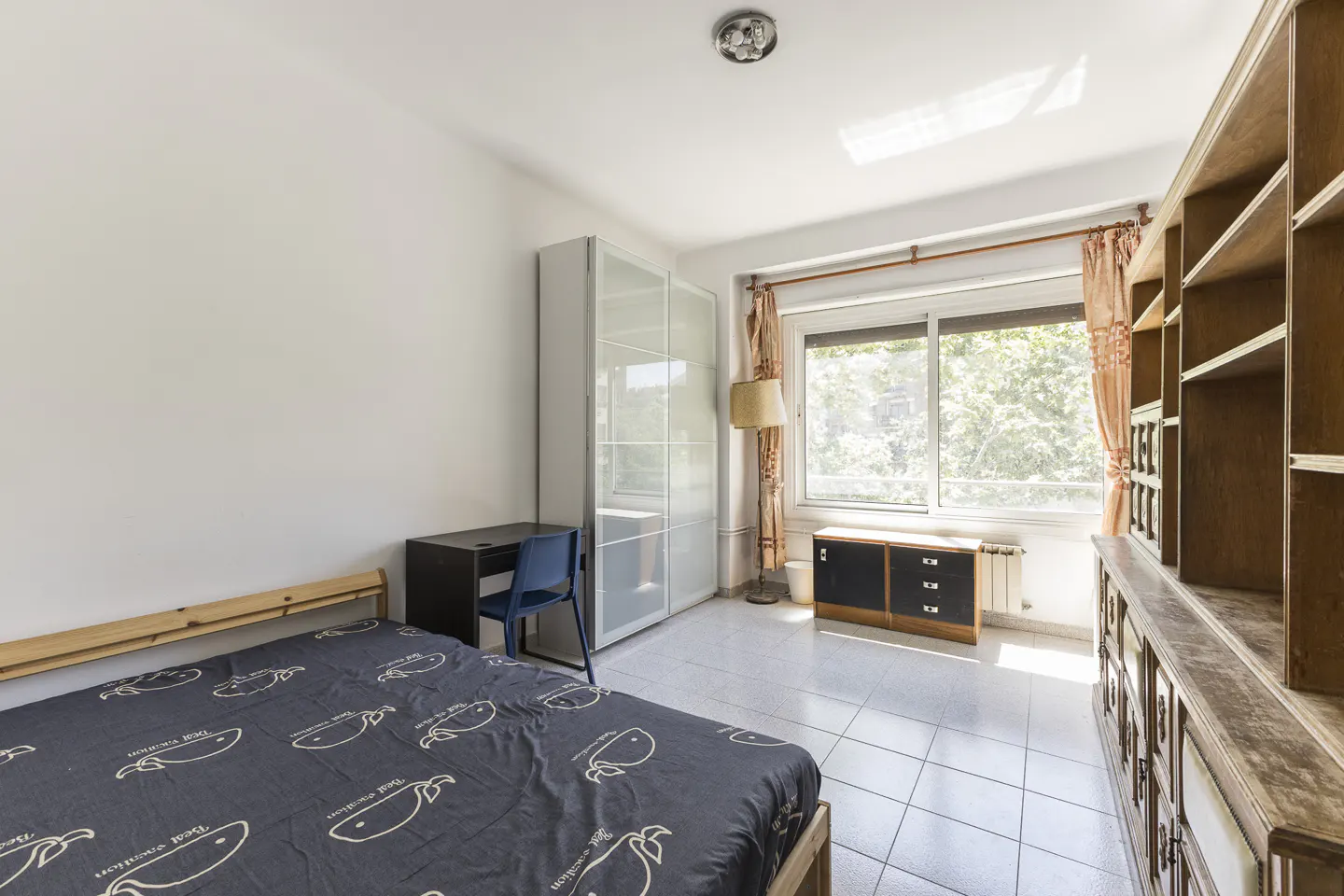 Bedroom with a bed, desk, blue chair, large wardrobe, and wooden shelving unit on a tiled floor near a bright window.