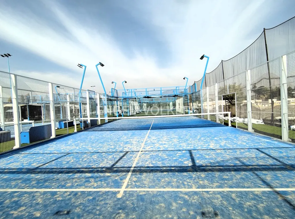 Outdoor blue paddle tennis court with white lines, net, and blue metal frame under a cloudy sky.