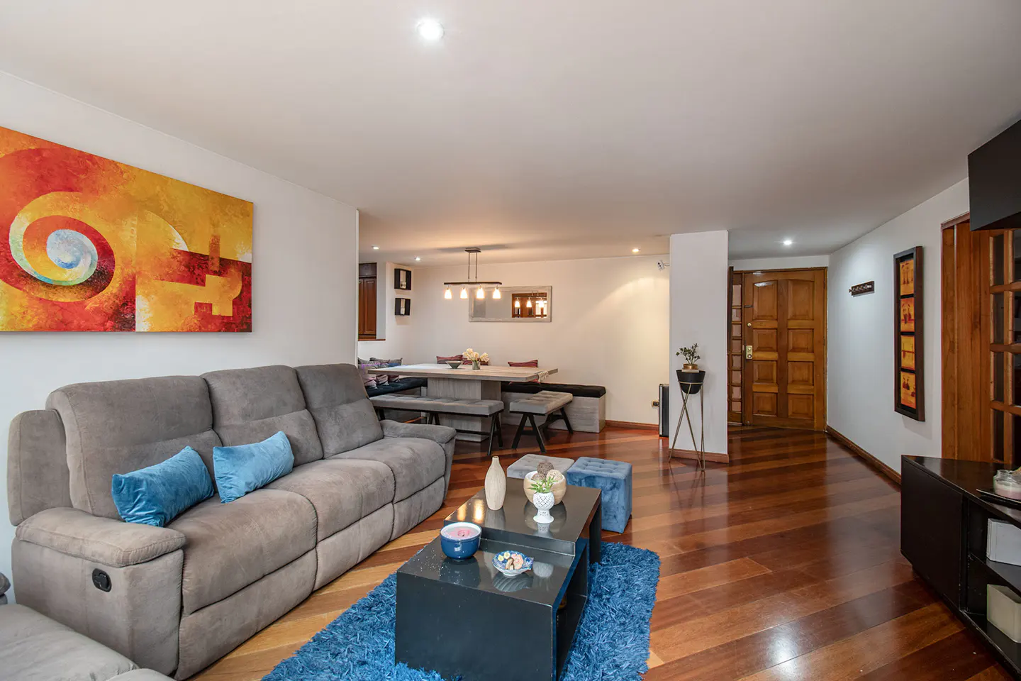 Living room with a gray sofa, blue pillows, and a black coffee table on a blue rug. A dining table is visible in the background.