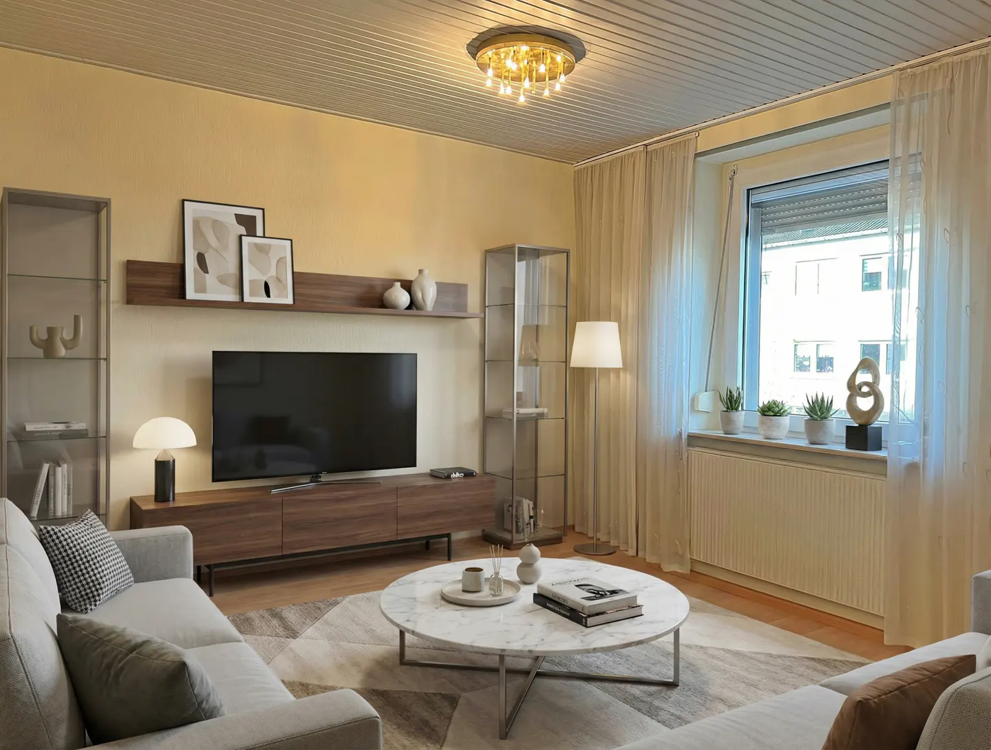 Living room with a gray sofa, marble coffee table, and a large TV. The walls are light yellow, and there's a window with white curtains.
