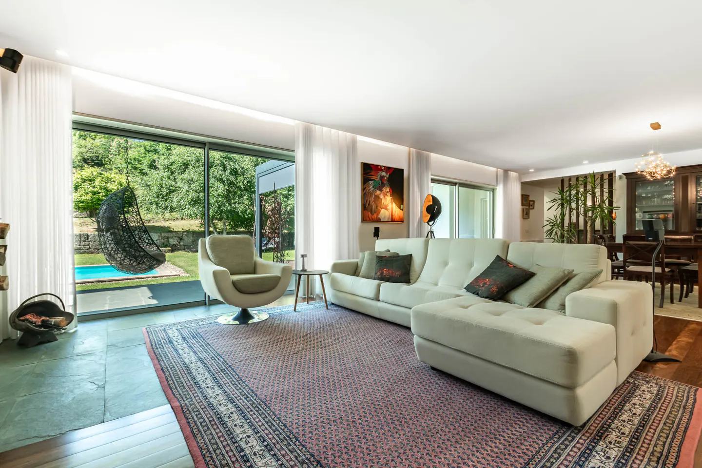 Bright living room with a white sofa, swivel chair, and a hanging chair visible through a large window overlooking a pool.