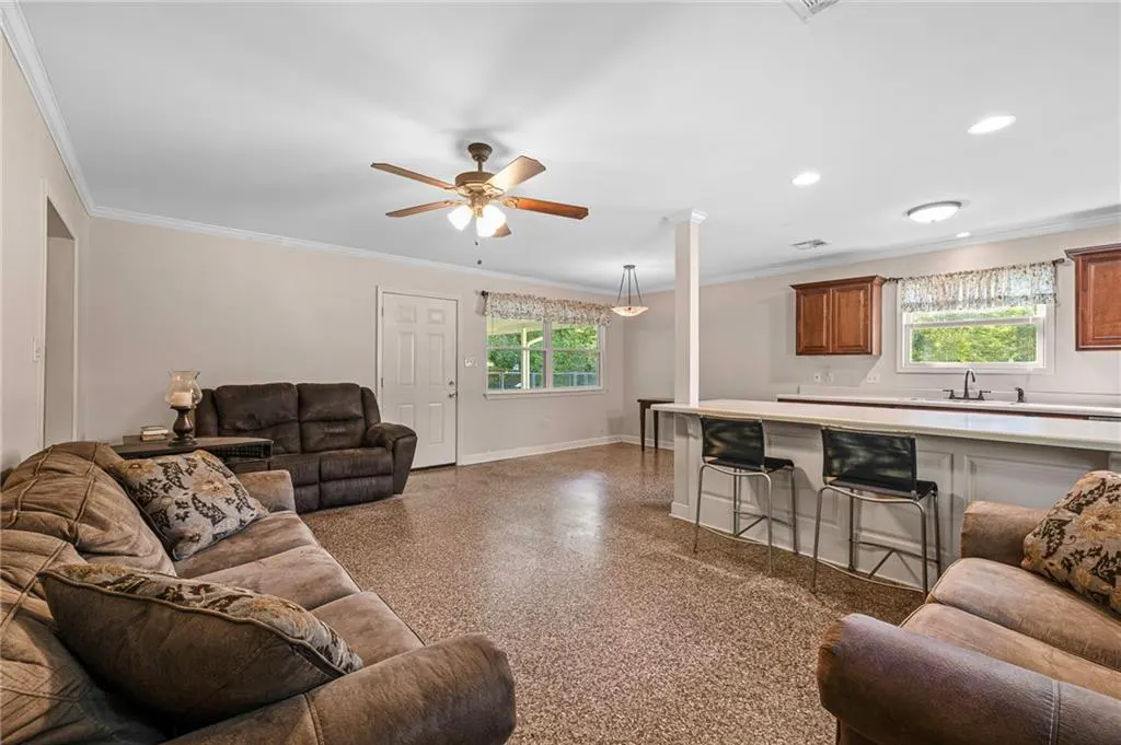 Open-concept living room and kitchen with brown couches, bar stools, and speckled flooring. A ceiling fan hangs above.