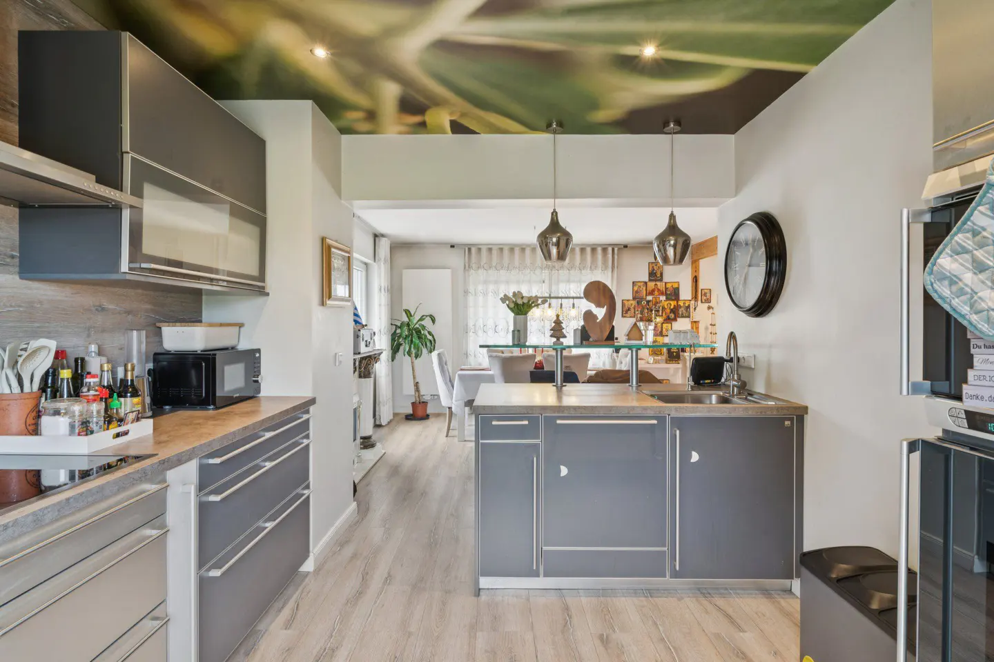 A modern kitchen with gray cabinets, light wood floors, and a unique ceiling mural of green leaves. Pendant lights hang above an island with a sink.