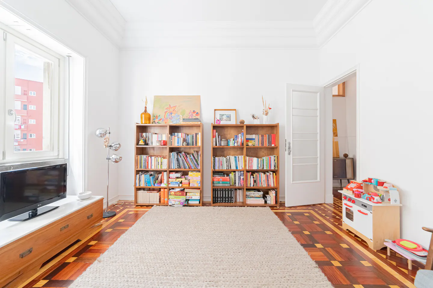 Bright living room with white walls, parquet floors, and a large beige rug. Two full bookshelves stand against the wall, and a toy kitchen sits near an open doorway.