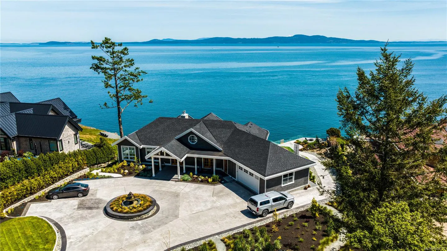Aerial view of a gray house with a dark roof, a circular driveway with a fountain, and a blue ocean backdrop.
