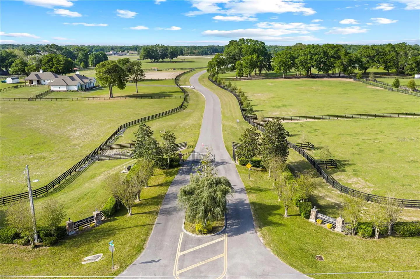 Aerial view of a long driveway leading to a house, surrounded by green fields and black fences.