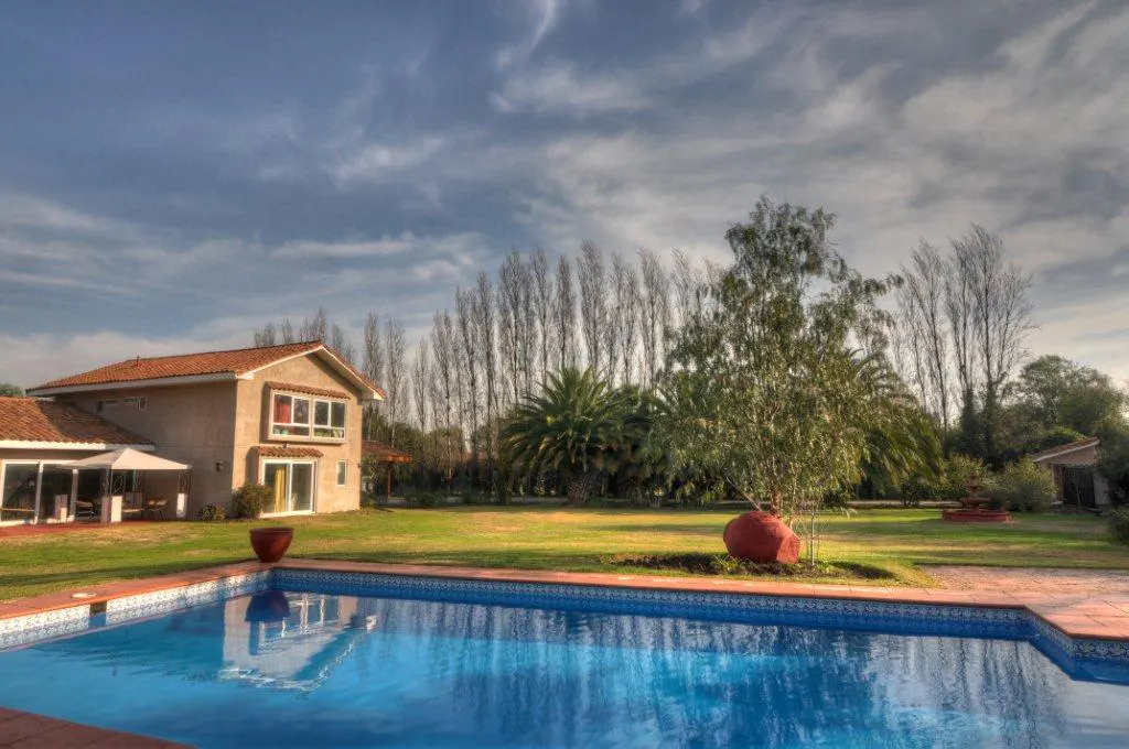 A backyard with a blue tiled pool, a tan house with a red tile roof, and green grass under a cloudy sky.