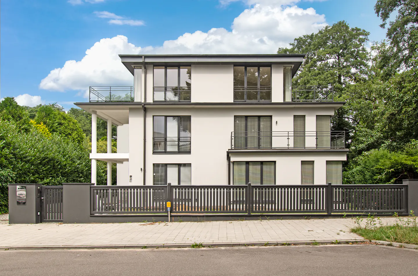 Three-story modern white house with black trim, balconies, and a gray fence, set against a blue sky with fluffy white clouds.