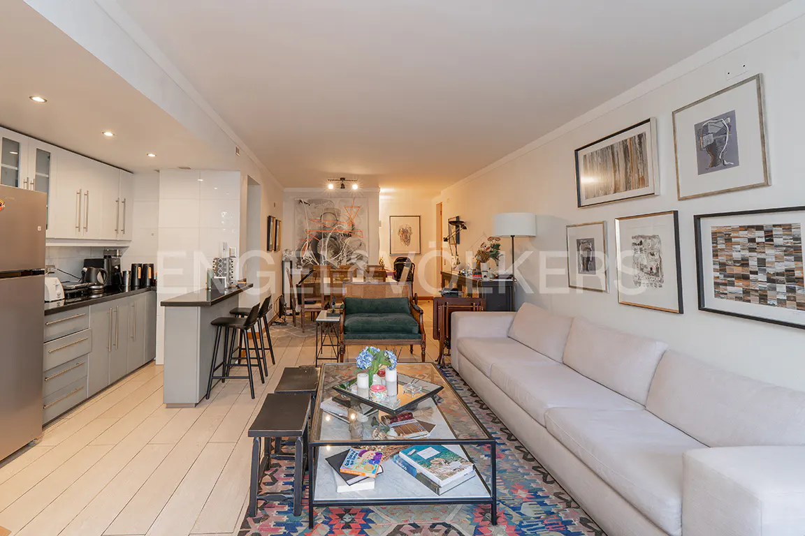 A wide shot of a living room with a white sofa, glass coffee table, and kitchen bar. Artwork adorns the walls.