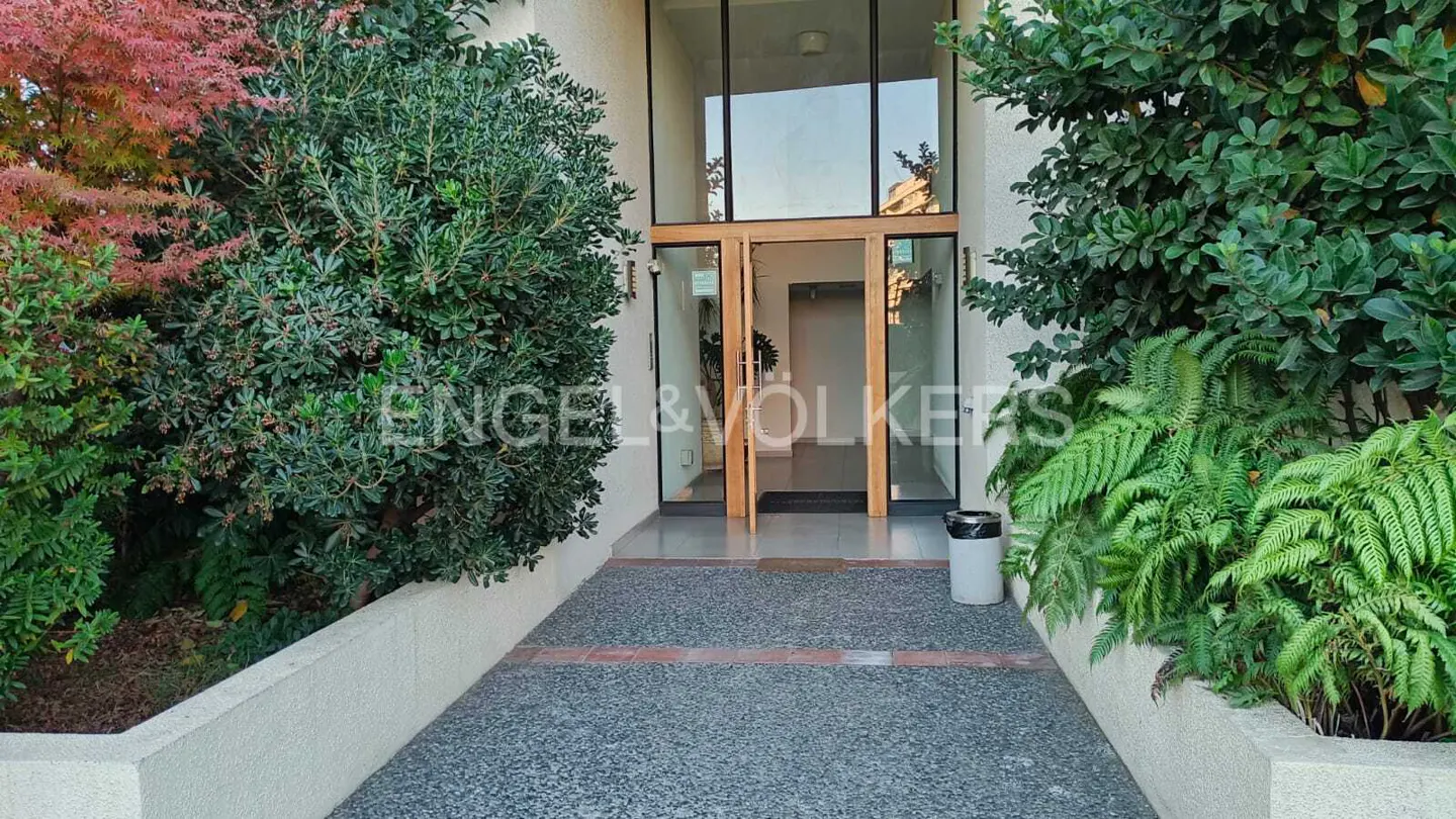 Exterior view of a building entrance with glass doors, surrounded by lush greenery and the Engel & Völkers logo.