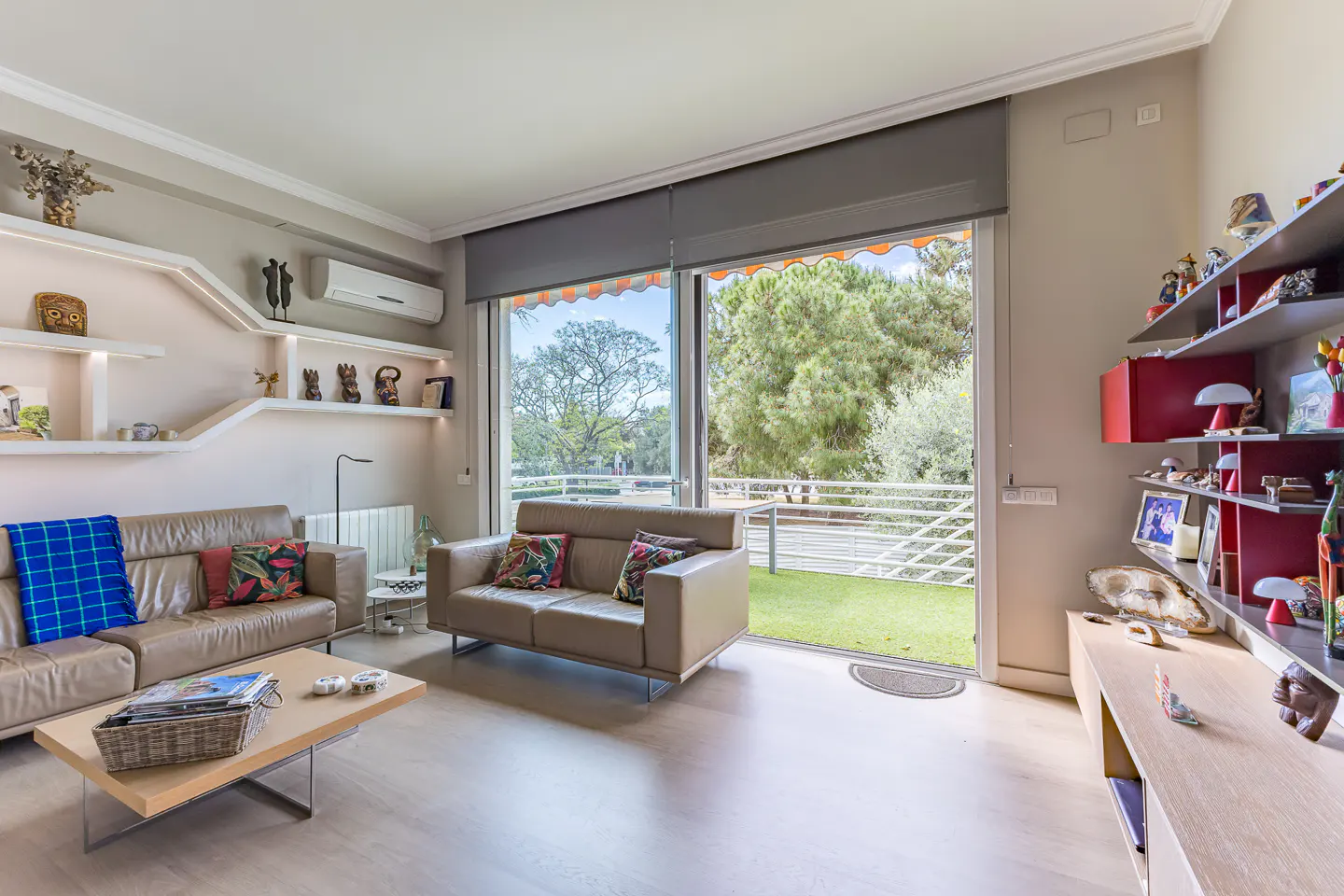 Bright living room with tan sofas, a light wood coffee table, and shelves with decorative items. Large windows overlook a green lawn and trees.