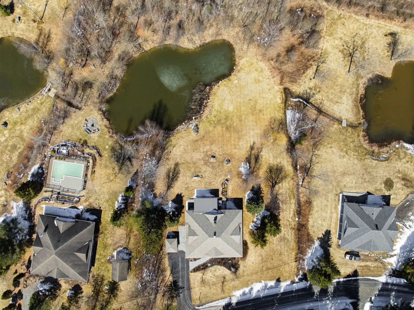 Aerial view of three houses with gray roofs, a pool, and three ponds on a brown, grassy lot with bare trees.