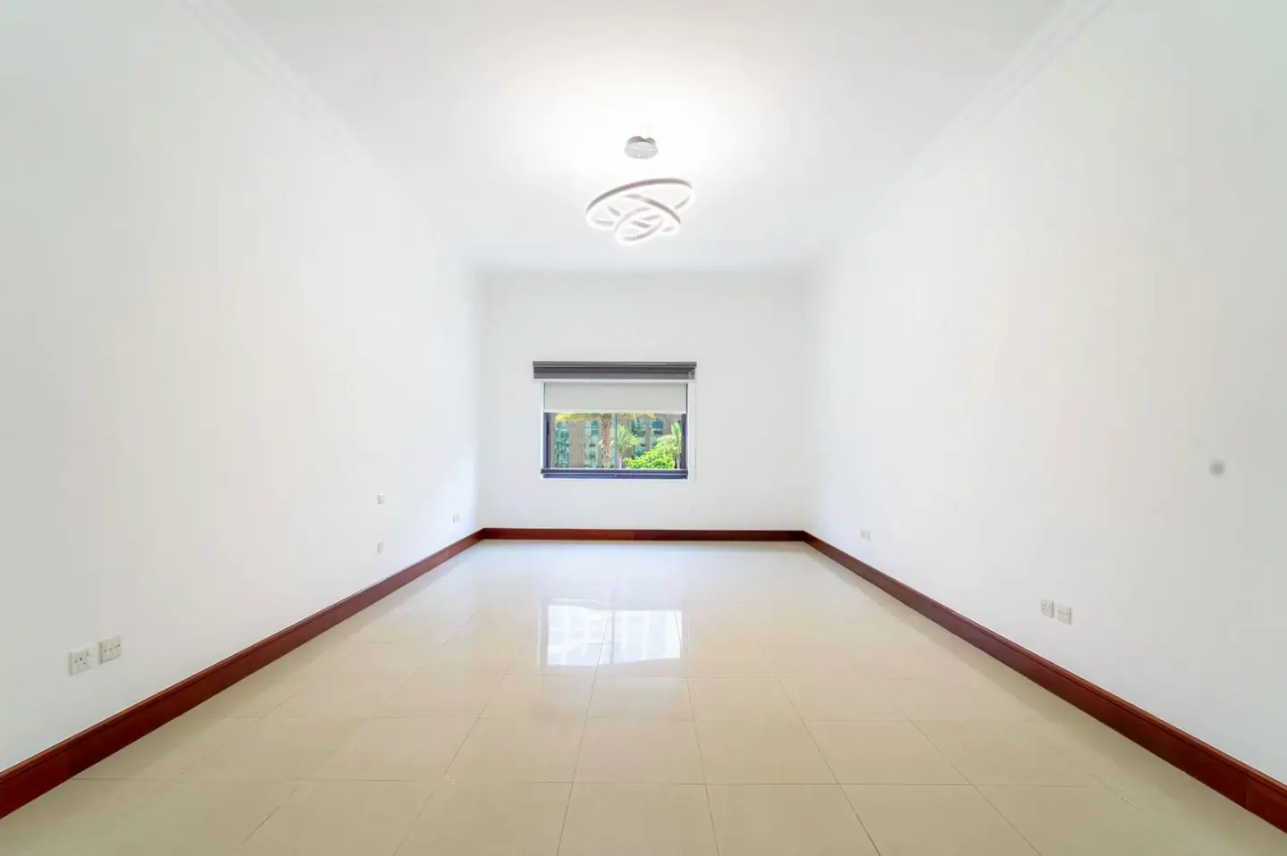 Bright, empty room with white walls, tile floor, and dark wood trim. A window with a shade looks out onto green trees. A modern light fixture hangs from the ceiling.