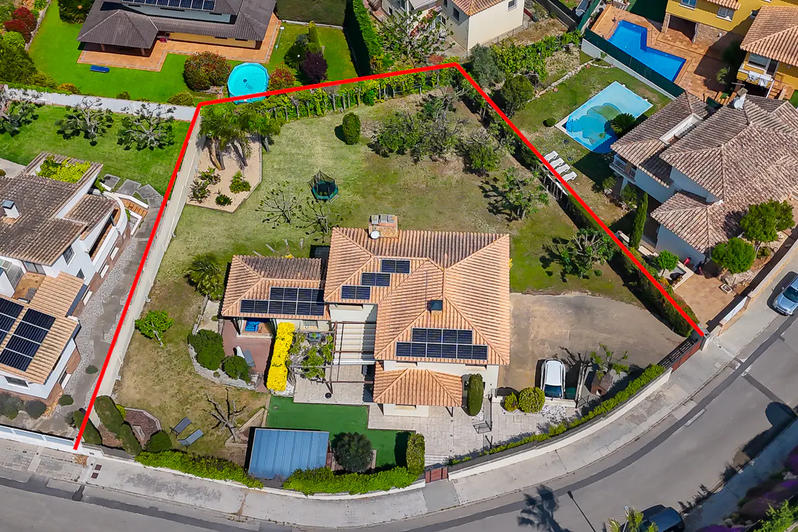 Aerial view of a tan house with a red tile roof and solar panels, surrounded by a green lawn and a red boundary line.