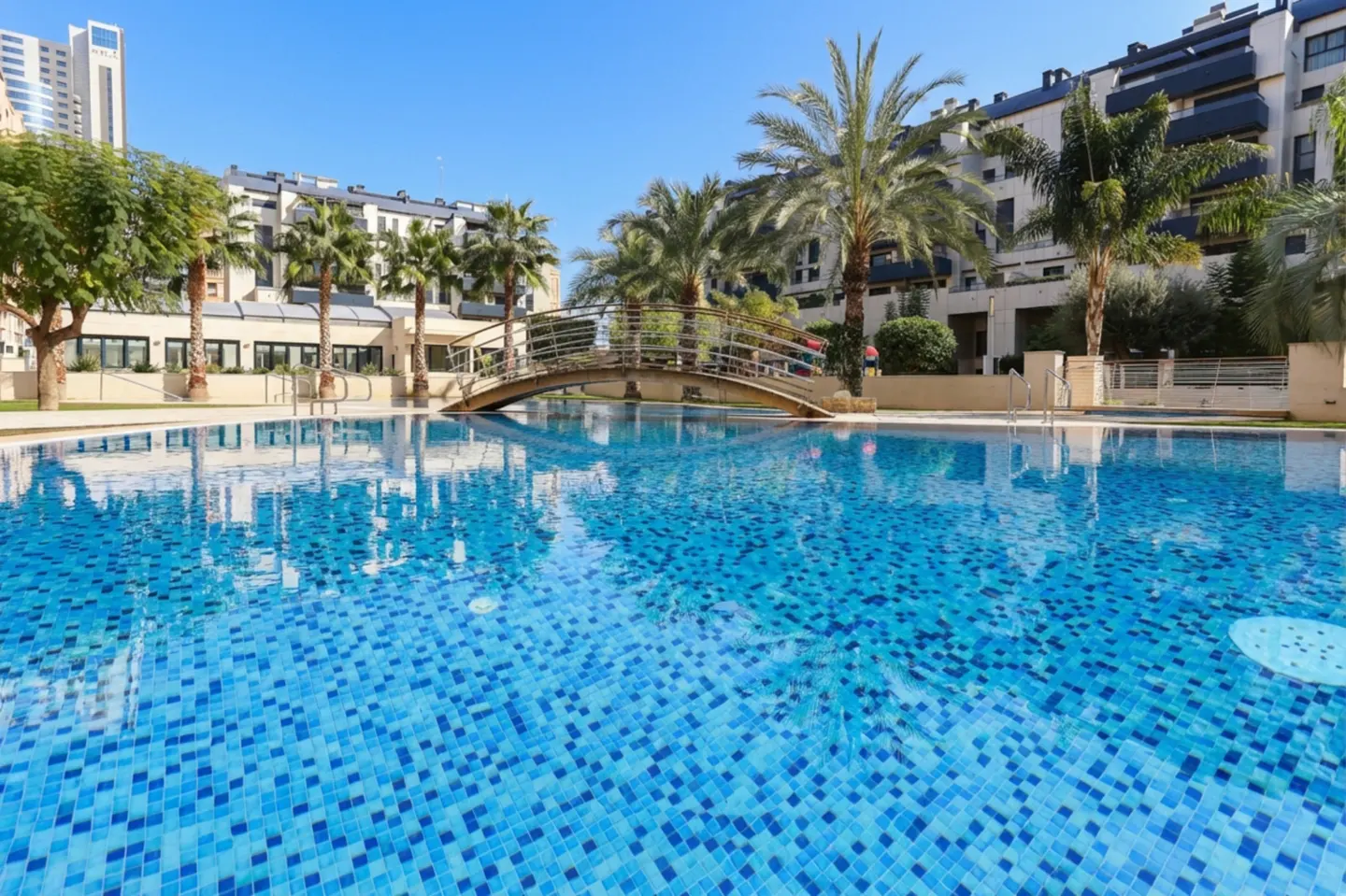 A blue tiled pool with a bridge over it, palm trees, and apartment buildings in the background.