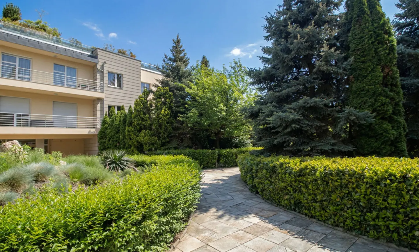 Stone path leads through manicured green hedges to a beige apartment building with balconies and tall trees under a blue sky.