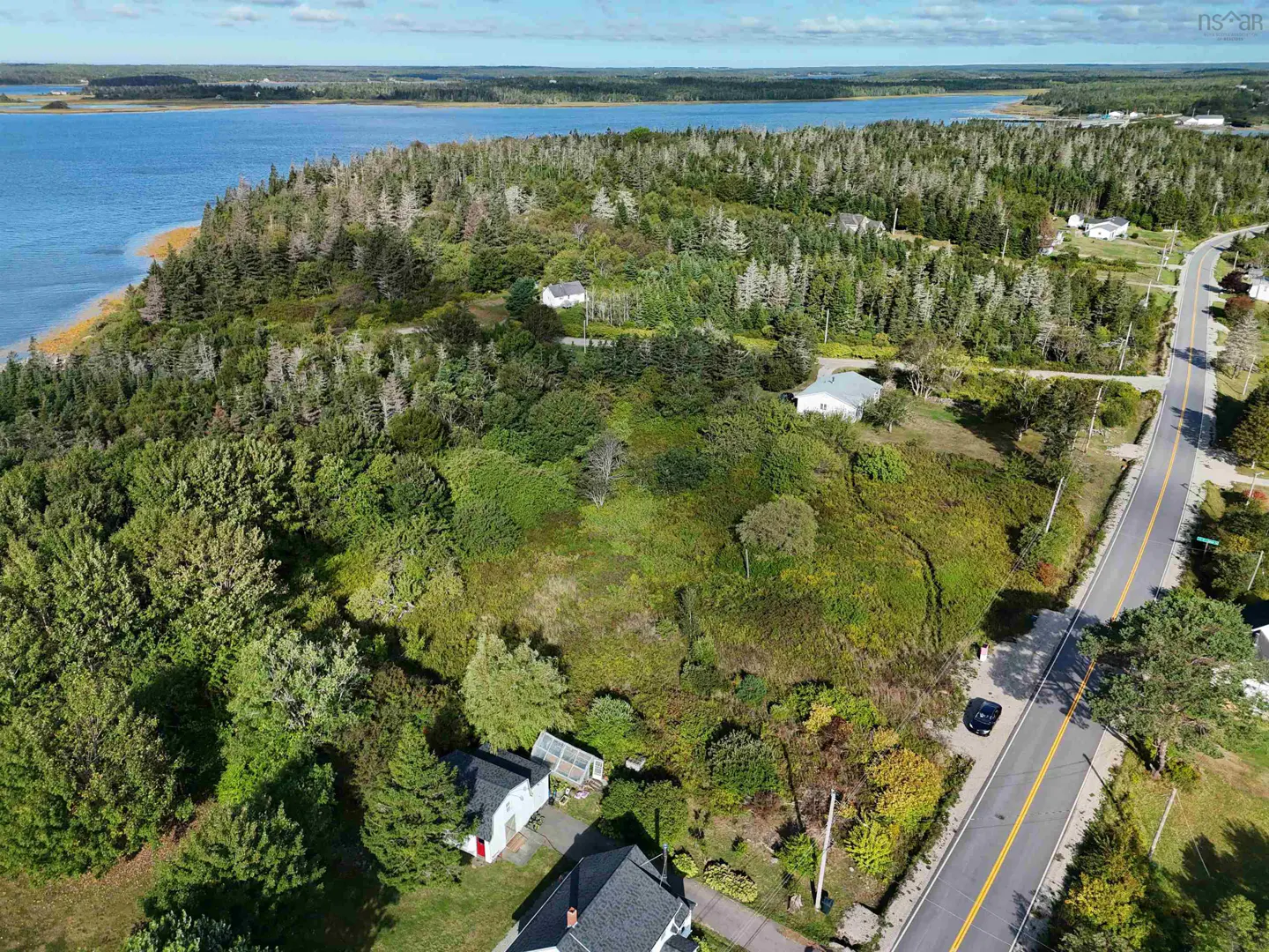 Aerial view of a coastal property with dense trees, a road, and small houses near a blue bay under a clear sky.
