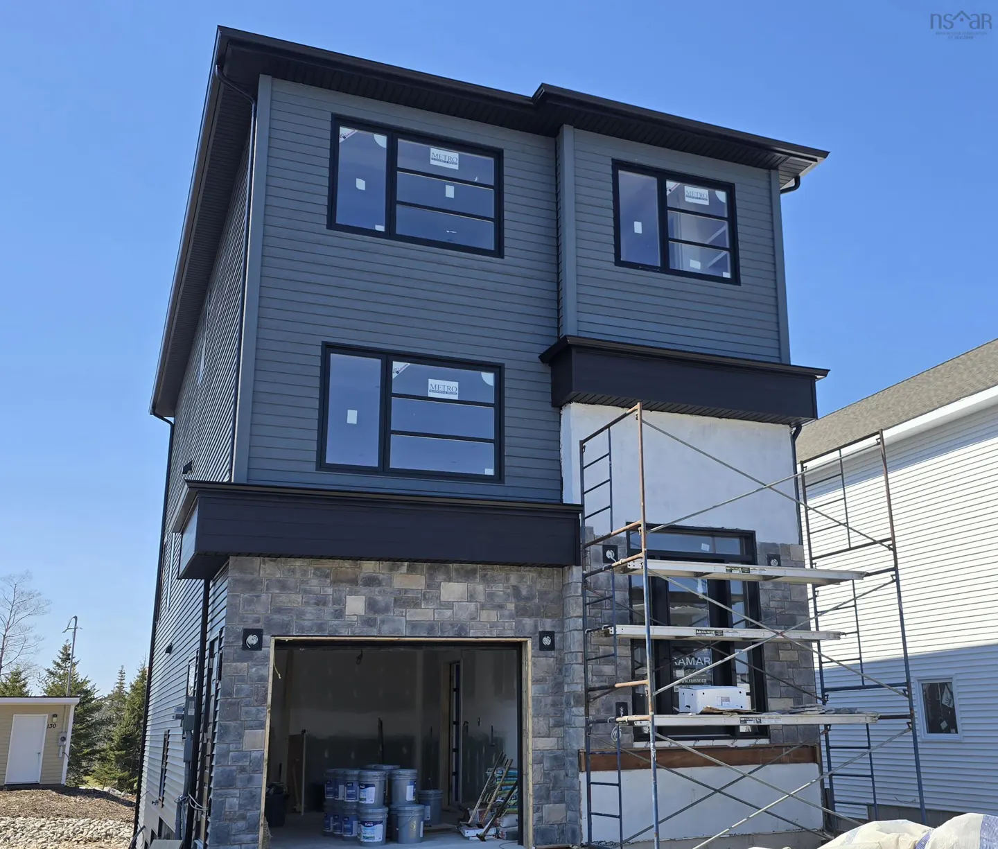 Two-story house with gray siding, black trim, and stone accents. Scaffolding is present, indicating ongoing construction. Blue sky background.