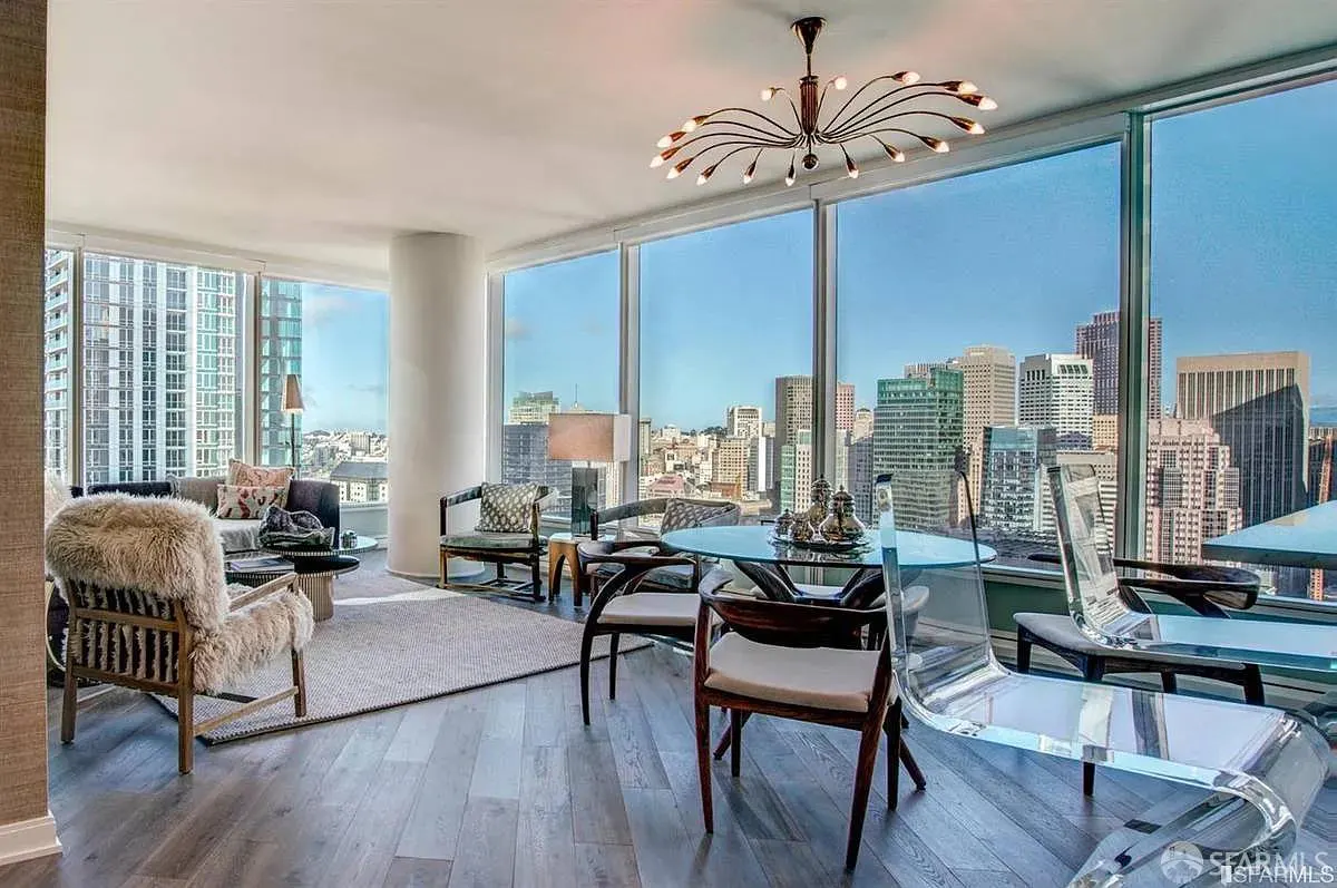 Bright living room with city view. Glass table, wood chairs, and a unique chandelier. Large windows showcase the urban skyline.