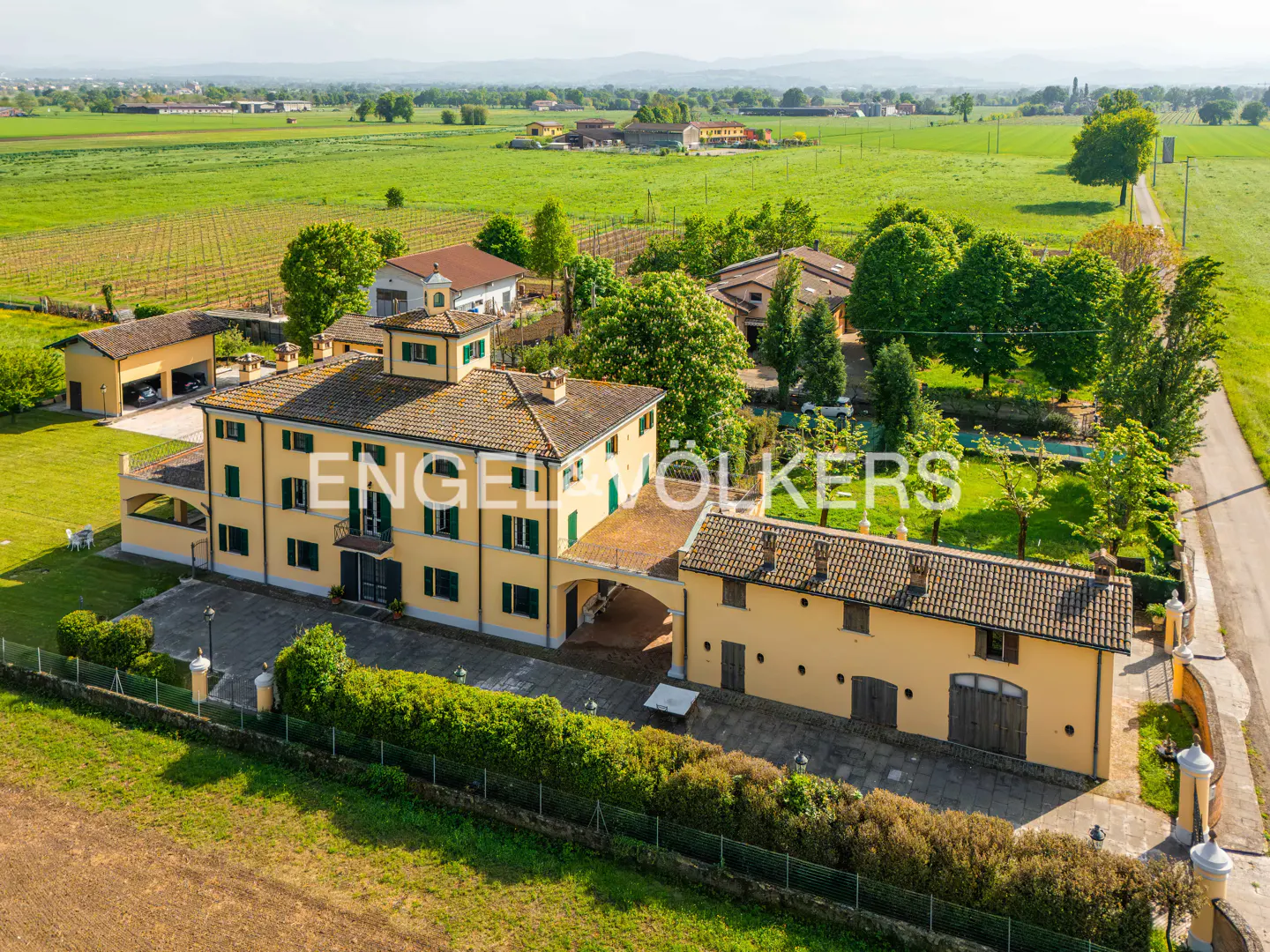 Aerial view of a yellow, two-story villa with green shutters, surrounded by green fields and trees. The Engel & Völkers logo is superimposed on the building.