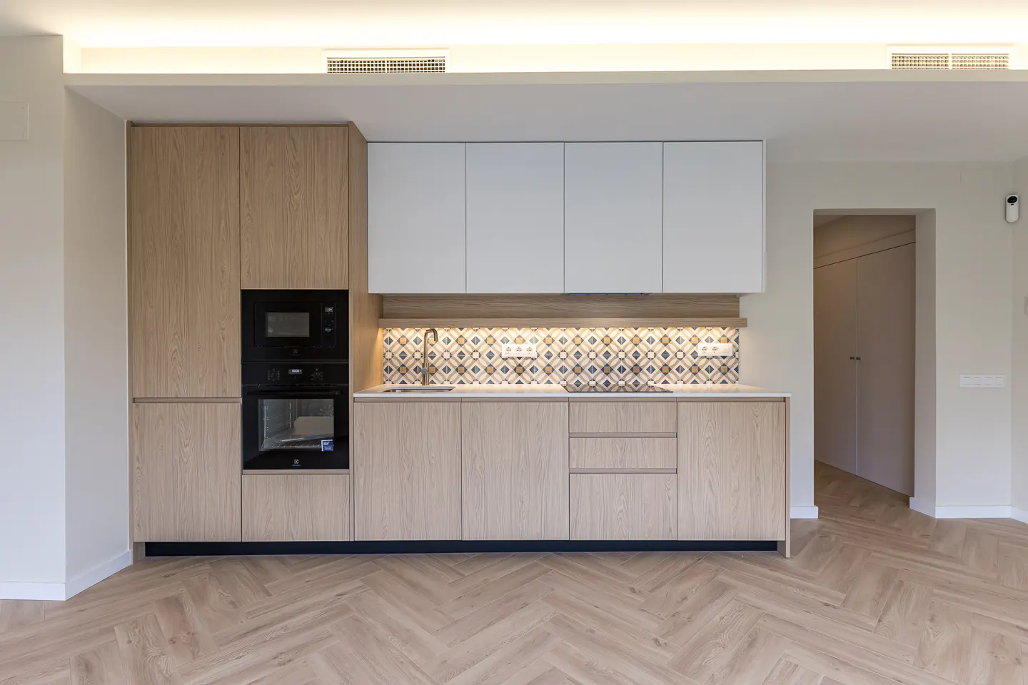 Modern kitchen with light wood cabinets, white upper cupboards, patterned tile backsplash, and herringbone wood flooring.