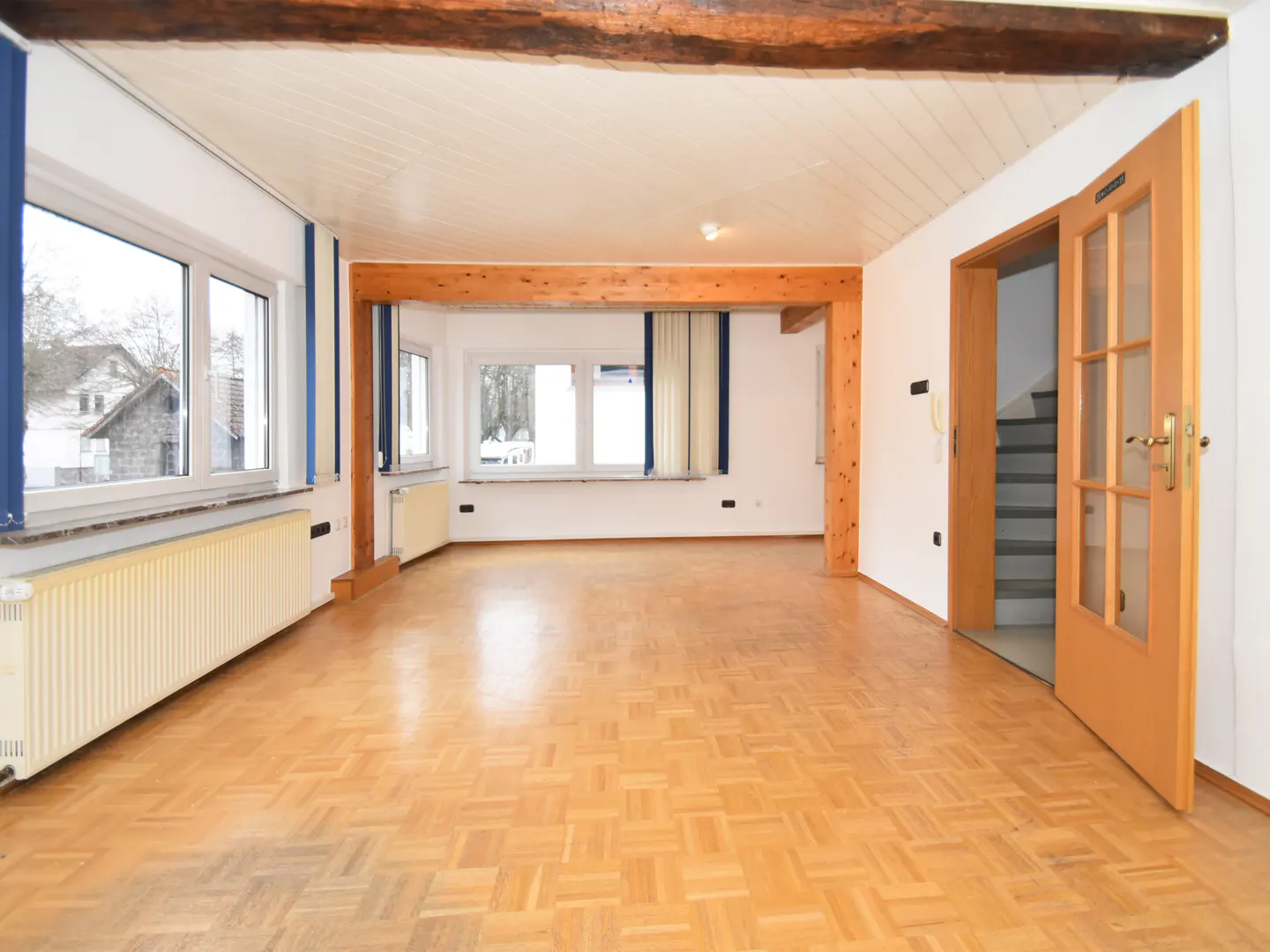 Bright, empty room with parquet flooring, white walls, and wooden beams. Windows show a view outside. A wooden door leads to stairs.