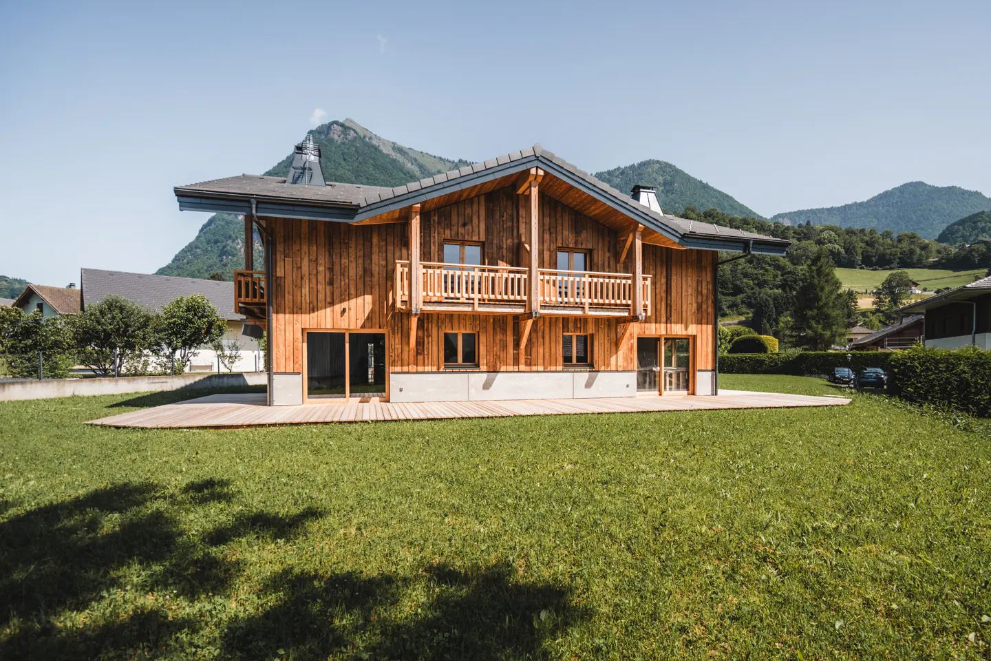A two-story wood chalet with a balcony, set on a green lawn with mountains in the background.