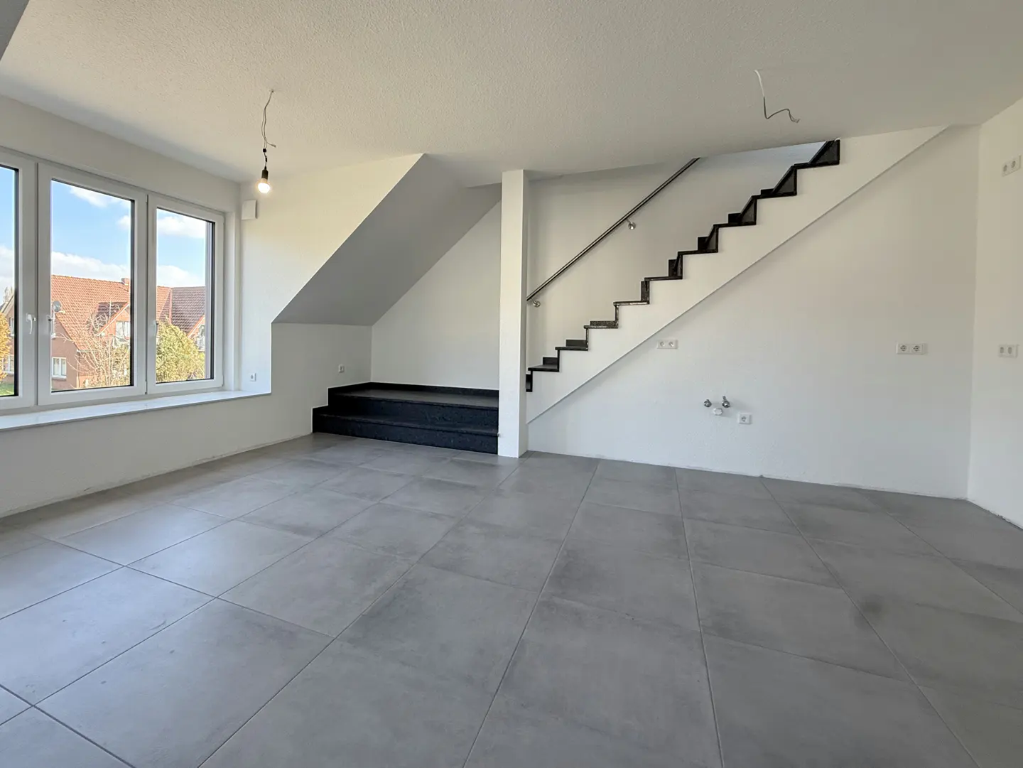 Bright, empty room with gray tile floor, white walls, and black stairs with a silver railing. Large windows show a neighborhood outside.