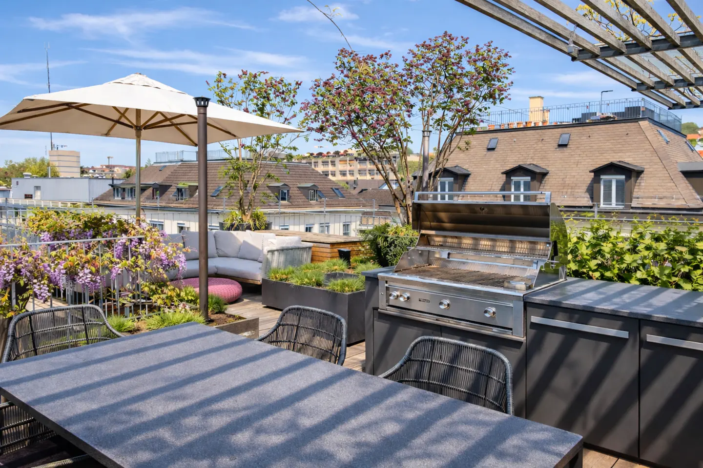 Rooftop patio with a table, chairs, grill, and lounge area. Flowers and greenery add color. City buildings are visible in the background.