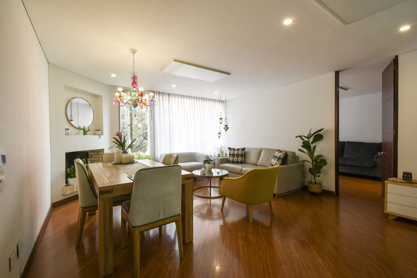 Bright, open-concept living and dining area with wood floors. A colorful chandelier hangs above a wooden dining table. A gray sofa and yellow chair sit in the living area.
