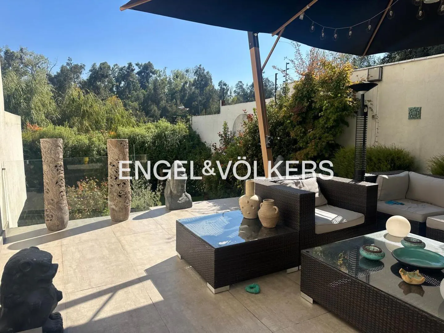 Outdoor patio with wicker furniture, glass tables, and decorative vases. Lush greenery and a black umbrella provide shade. Engel & Völkers logo visible.