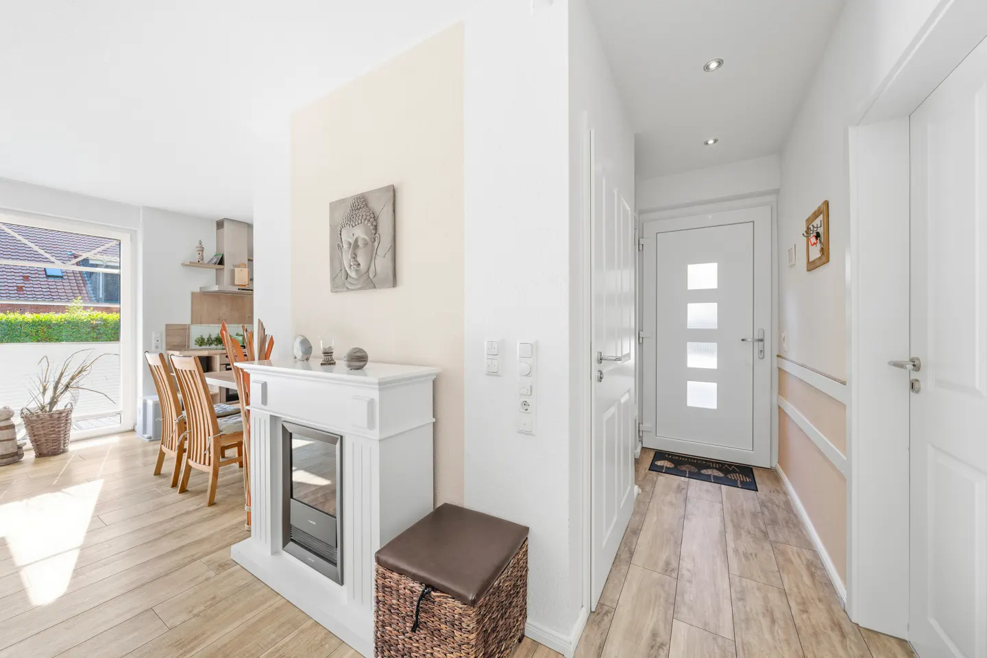 Bright entryway with white walls, light wood floors, and a white front door with four glass panels. A white fireplace and Buddha art add character.