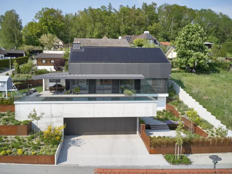 Modern gray house with solar panels, a pool, and a garage. The house is surrounded by greenery and other houses.
