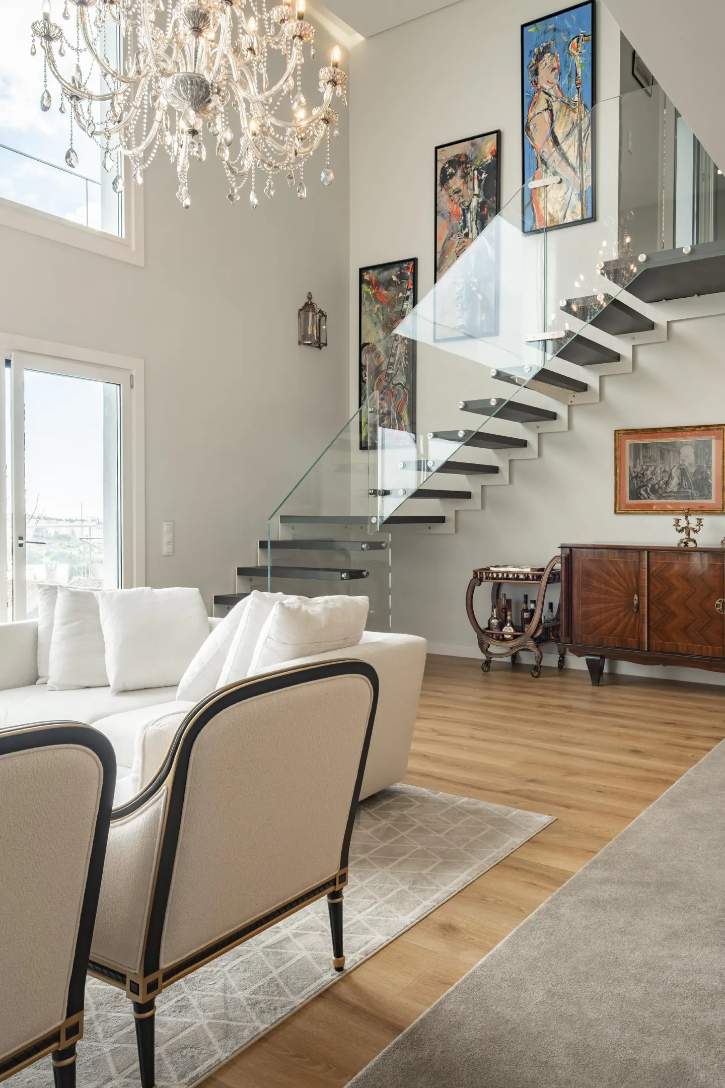 Modern living room with white sofas, a crystal chandelier, and a floating staircase with glass railings and wall art.