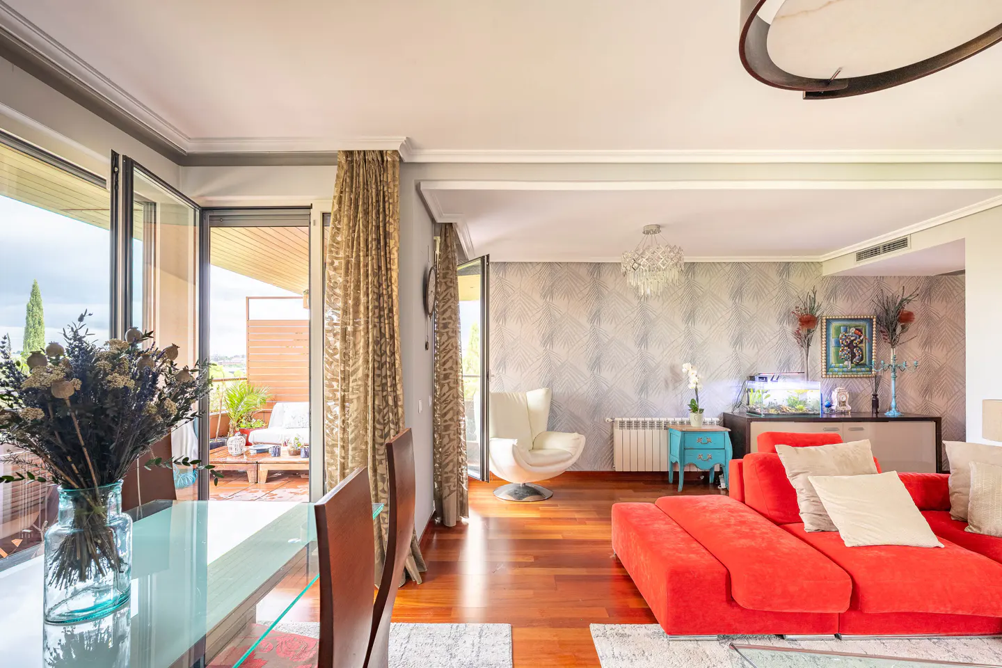 Bright living room with hardwood floors, red sofa, glass table with flowers, and balcony access.