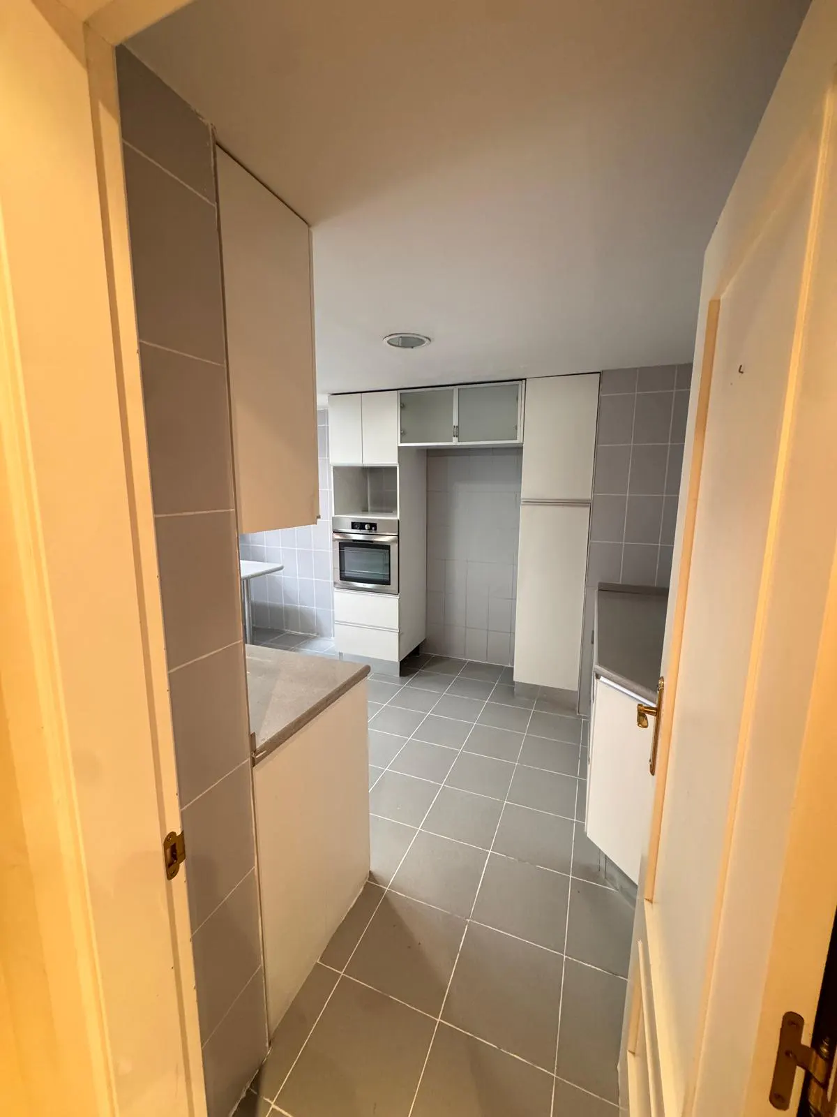 A bright kitchen with gray tile floors and white cabinets, viewed from a doorway. A stainless steel oven is built into the cabinets.