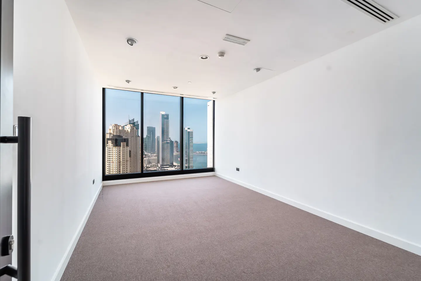 Empty room with beige carpet, white walls, and a large window showing a city skyline and ocean view.