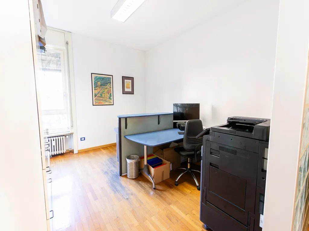 Office space with a blue desk, black chair, computer, and printer. Hardwood floors and white walls. Two framed pictures hang on the wall.