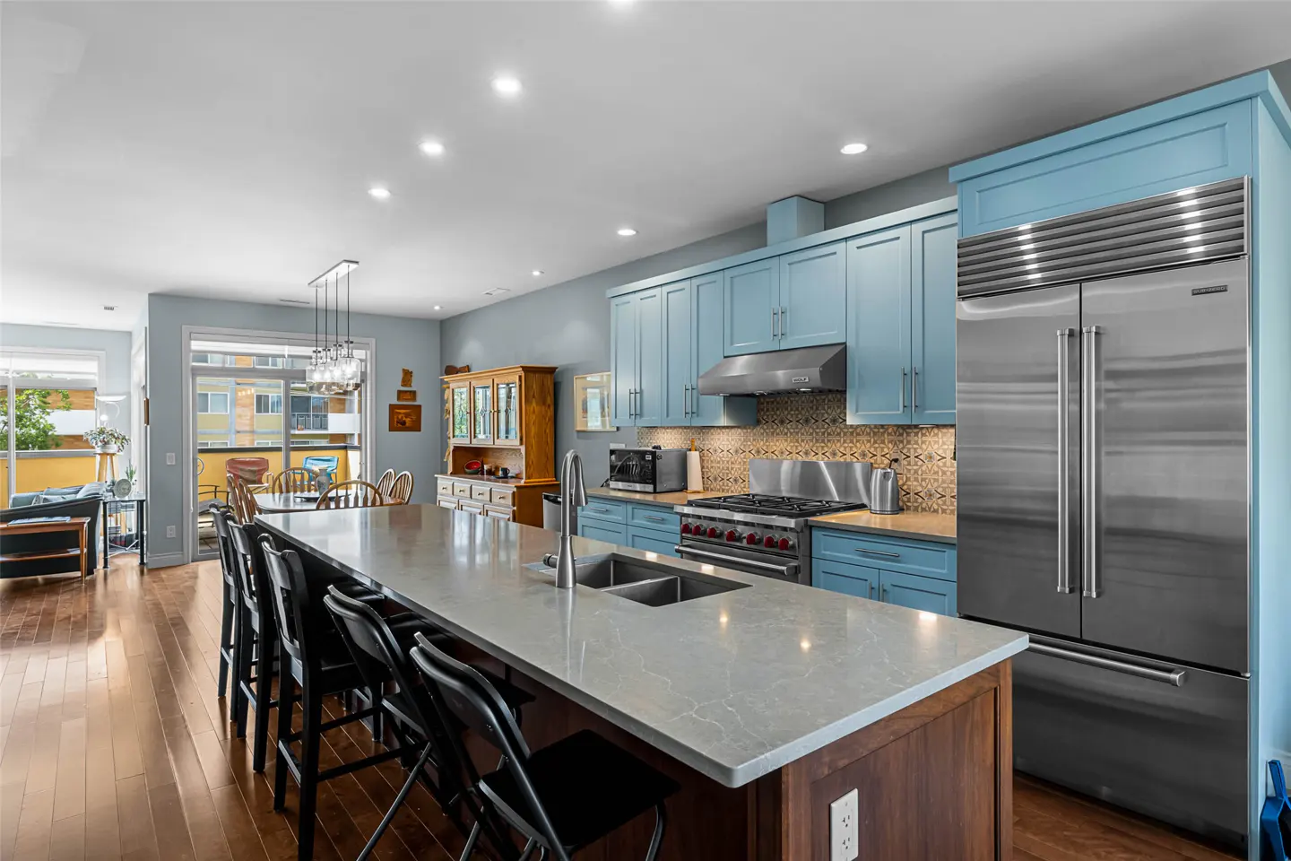A modern kitchen with blue cabinets, stainless steel appliances, and a large island with black stools. Hardwood floors and a dining area are visible.