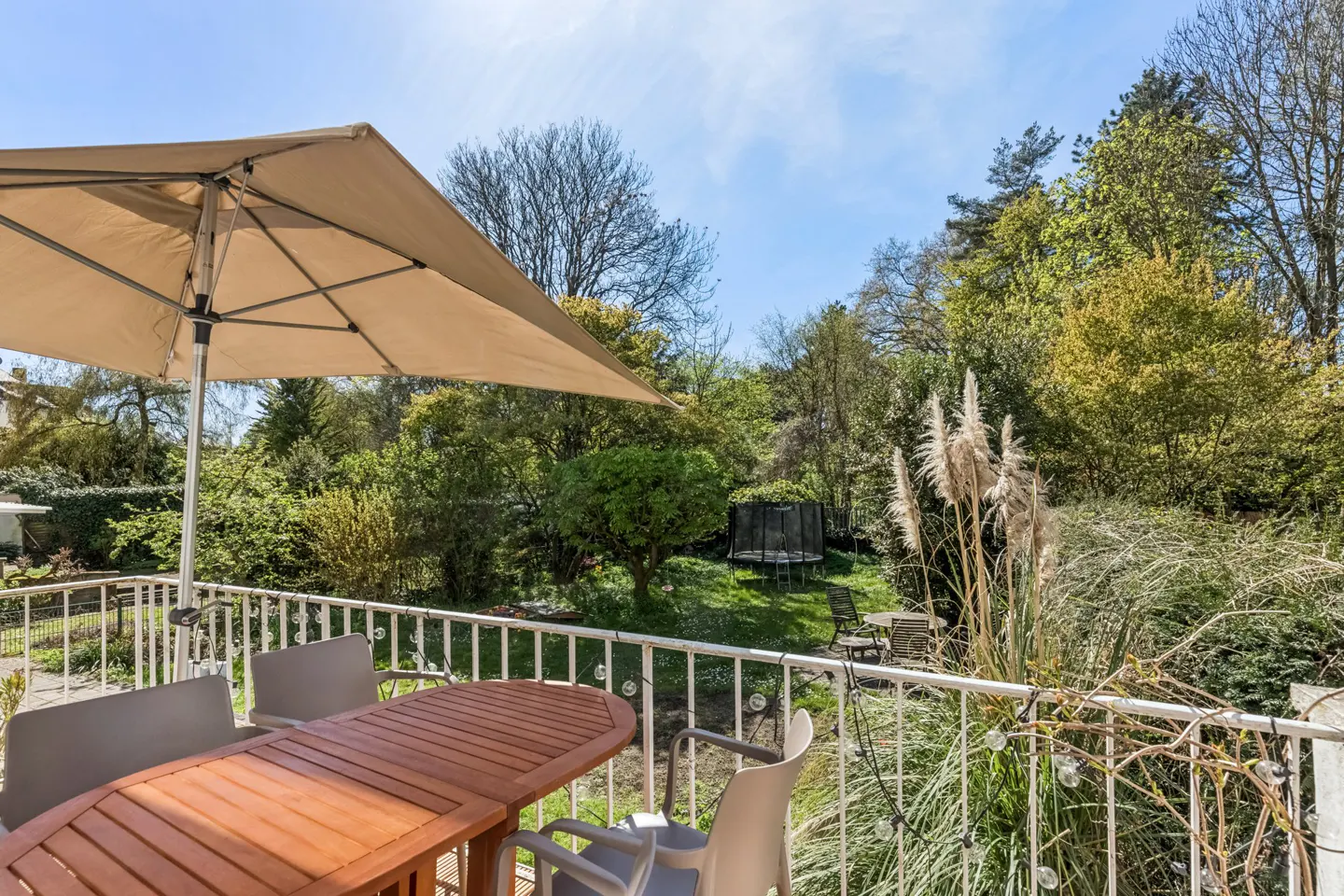 Balcony view with a brown wooden table, gray chairs, and a beige umbrella. A green garden with a trampoline is in the background.
