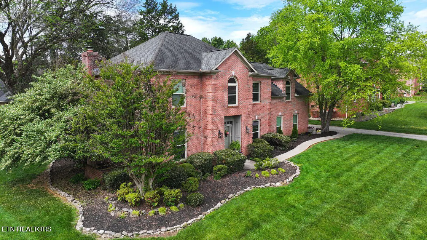Two-story red brick house with a gray roof, surrounded by green trees and a well-manicured lawn.