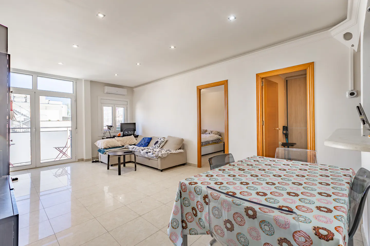 Bright living room with white walls, tile floor, and donut-patterned tablecloth on a table with transparent chairs. A beige sofa and balcony are visible.