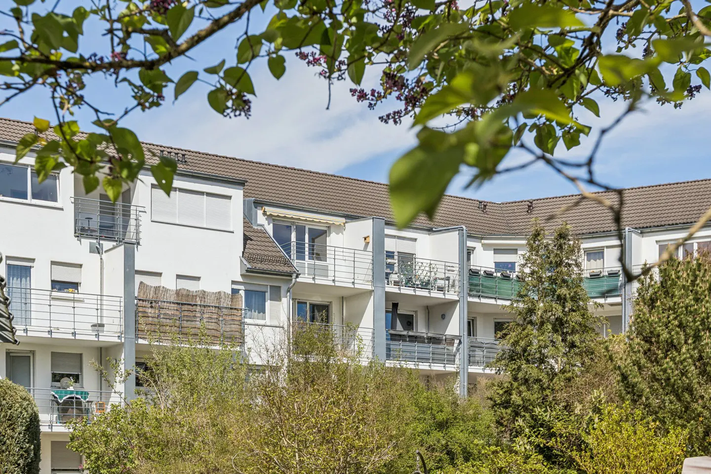 Exterior view of a white apartment building with balconies, brown roof, and green trees in the foreground.