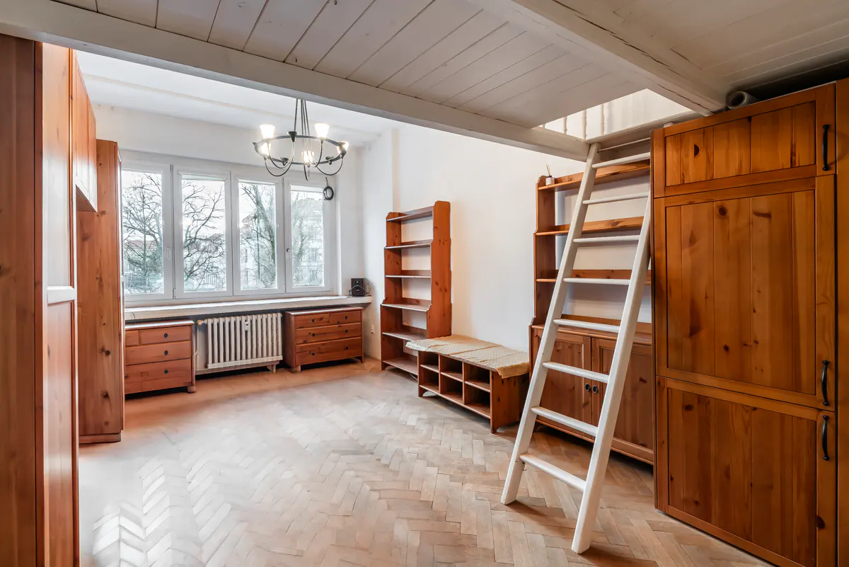 Bright room with wood floors, white walls, and wood furniture. A white ladder leads to a loft area. A window overlooks trees.