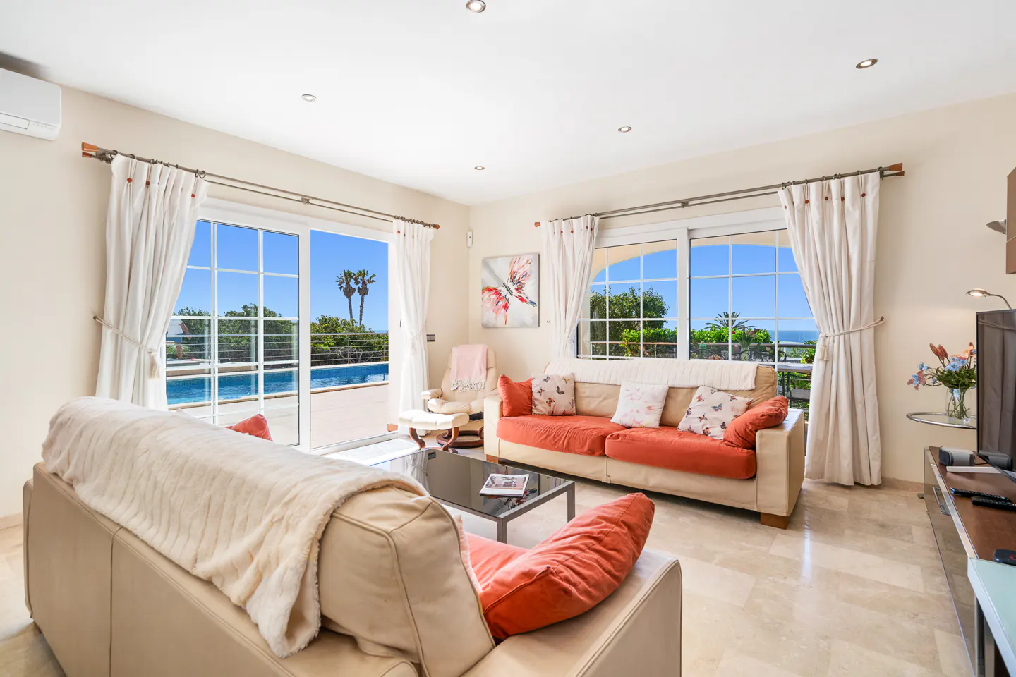 Bright living room with beige sofas, orange cushions, and a glass coffee table. Sliding glass doors open to a pool and palm trees.