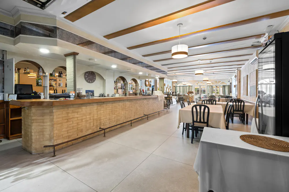 Interior view of a restaurant with a long bar, tables with white tablecloths, and wooden beams on the ceiling.