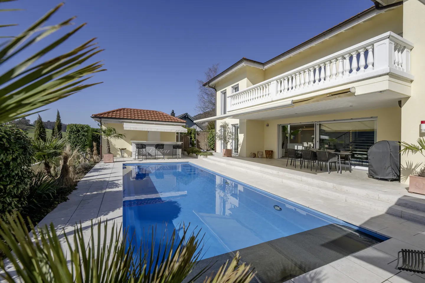 A sunny backyard with a blue swimming pool, a bar, and a yellow two-story house.